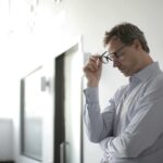 Thoughtful man in a bright room holding his glasses while leaning against a wall.