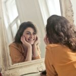 A cheerful woman smiles at her reflection in a vintage-style mirror, exuding positivity and warmth.